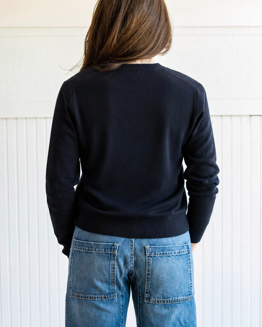 Person wearing a black sweater and blue jeans standing against a white paneled wall.