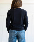 Person wearing a black sweater and blue jeans standing against a white paneled wall.